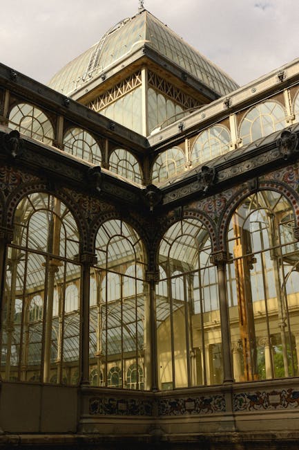 A detailed view of an ornate glass conservatory featuring intricate metal framework and large arched windows, with a portion of a historic building visible at the top, set against a cloudy sky. Inside the conservatory, several cardboard boxes and wrapped furniture items are placed on the floor, indicating preparations for home relocation or furniture transport. Part of a moving trolley and a van may be seen through the windows, suggesting the loading process for a house removal by [COMPANY_NAME]. The scene reflects the careful packing and loading activities associated with professional removals, with a focus on glass and metal architecture typical of historic properties often involved in house moves in the Crystal Palace area.