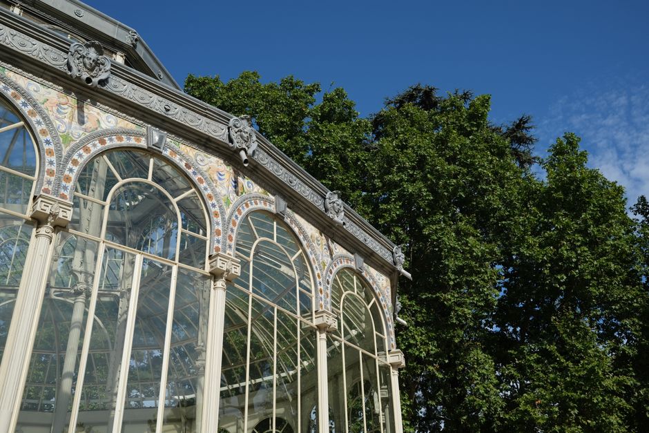 A large glasshouse building with a domed central section and extending wings, situated in a park surrounded by trees with autumn foliage in shades of orange, yellow, and green. The building features a